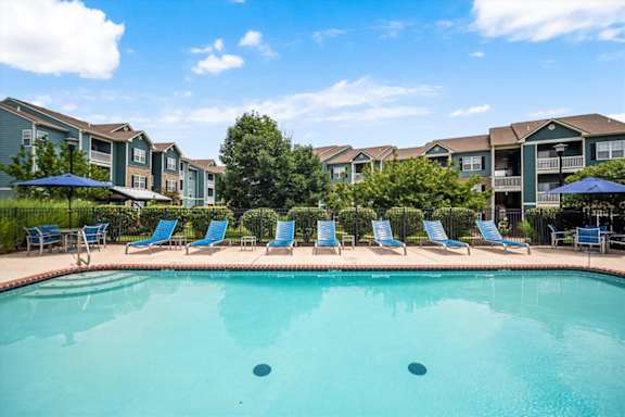 A swimming pool surrounded by blue lounge chairs and umbrellas in front of apartment buildings.
