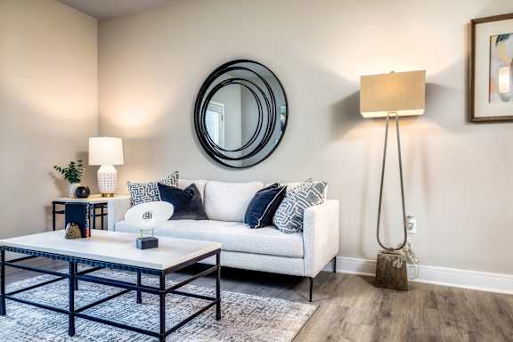A modern living room with a white sofa, a round mirror, and a wooden floor lamp.