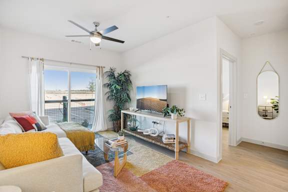 A living room with a white couch, a yellow throw pillow, a TV, and a large window with a view of the outdoors. at ThreeTwenty at the Vybe Apartments, Texas