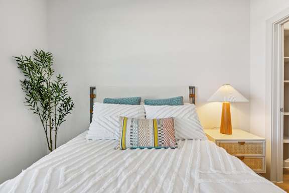 A bed with a white comforter and striped pillows is in a room with a lamp and a plant. at ThreeTwenty at the Vybe Apartments, Kyle, Texas