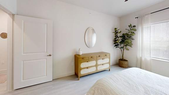 A white bedroom with a bed, dresser, mirror, and plant.