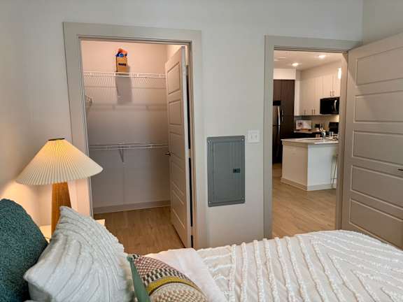 A bedroom with a bed, lamp, and a view into a kitchen.