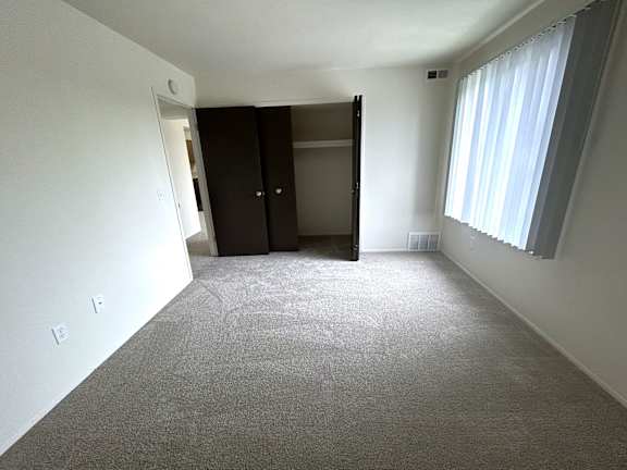 A bedroom with a carpeted floor, a closet with a door open, and a window with curtains.