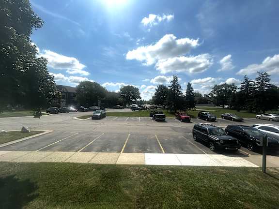A sunny view of grass and a parking lot with cars parked and trees in the background.