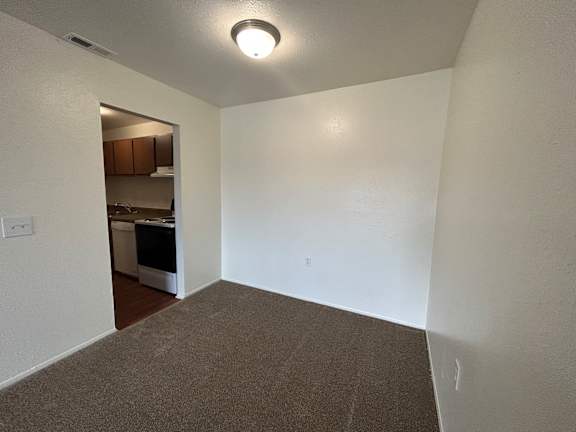 A dining room with a carpeted floor and a small kitchen visible through the door.