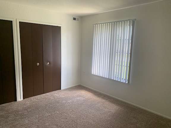 A bedroom with a carpeted floor, a window with blinds, and a brown wardrobe.