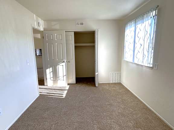 A bedroom with a carpeted floor, a window with a patterned curtain, and a door leading to another room.