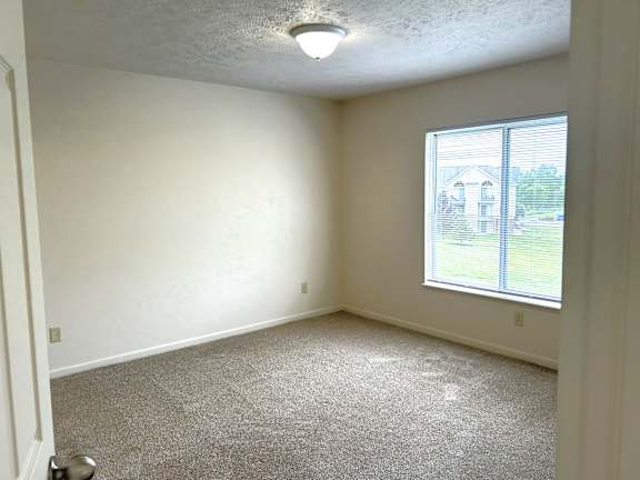 A bedroom with a carpeted floor and a window showing a view of a house.