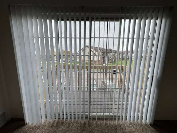 A view of green space and a building through a window with white blinds.