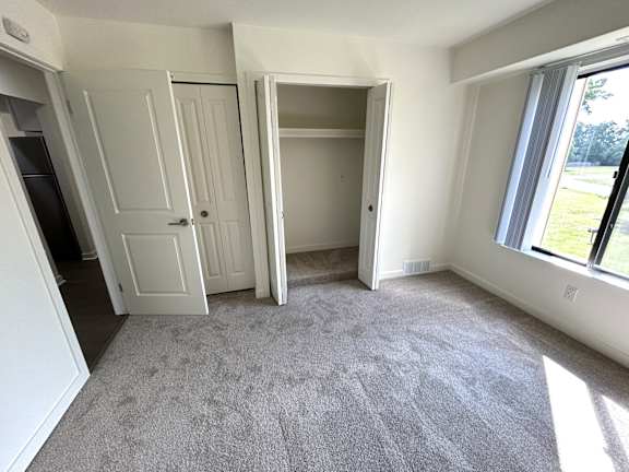 A bedroom with a carpeted floor and a window showing a green outdoor area.