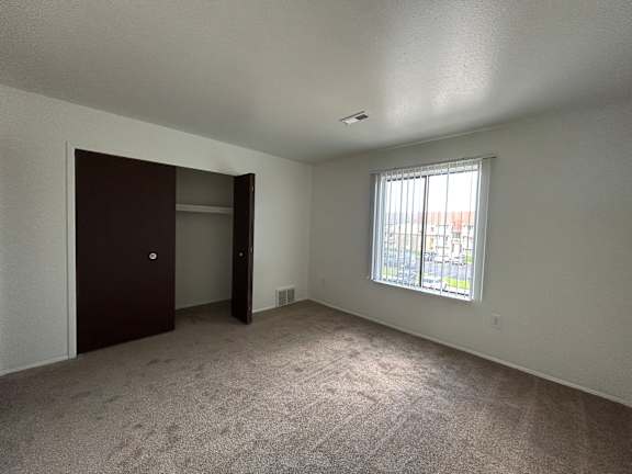 A bedroom with a carpeted floor, a window with blinds, and a brown door.