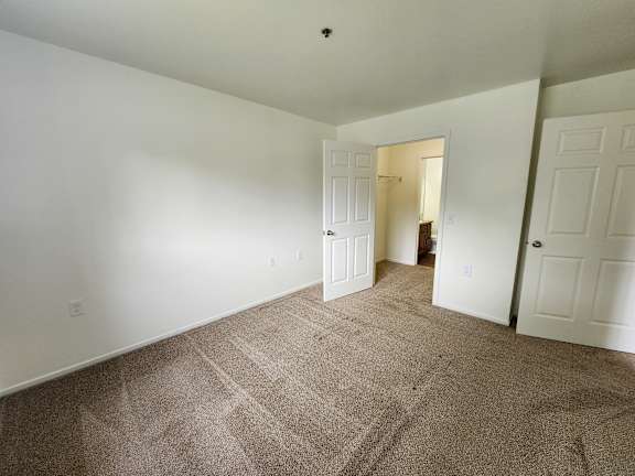 A bedroom with white walls and carpeted floor with a doorway leading to another room.
