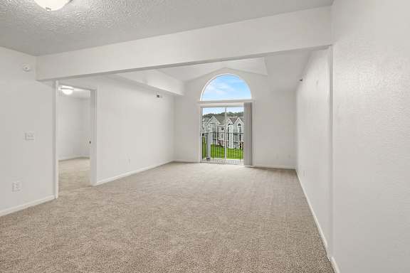 A spacious living room with a cathedral ceiling, carpeted floor and a view of a balcony and sky through an arched window at Autumn Lakes Apartments and Townhomes in Mishawaka, IN