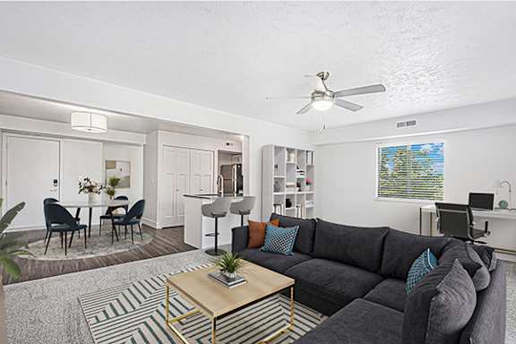 An enlarged living room with a grey couch, a coffee table, and a dining table with chairs at Byron Lakes Apartments in Byron Center, MI