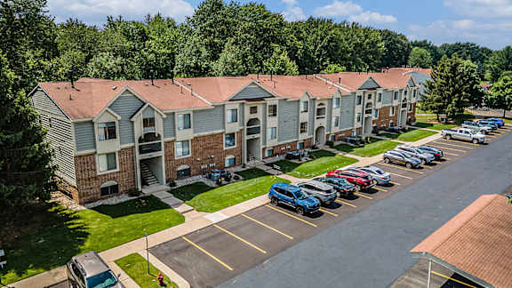 A parking lot in front of an apartment building with tall trees at Byron Lakes Apartments, Byron Center, Michigan
