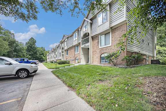 An exterior of an apartment building with a sidewalk and shade trees at Byron Lakes Apartments, Byron Center, Michigan