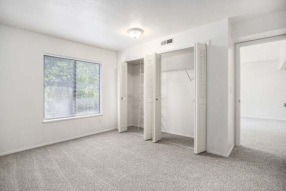 A bedroom with white walls and a carpeted floor with a window and open closet doors at Canal Club Apartments in Lansing, MI