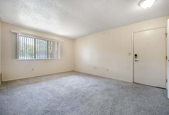A garden style living room with a carpeted floor, a window with blinds, and a door at Concord Place Apartments, Kalamazoo, MI, 49009