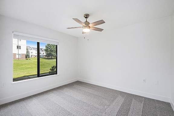 A bedroom with a ceiling fan and a window overlooking a green lawn at The Crossings Apartments in Grand Rapids, MI