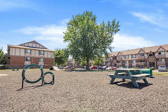 A playground with a green bench and a tree in the foreground and apartment buildings in the background at Pheasant Run Apartments, Indiana, 47909