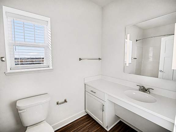 A white bathroom with a toilet, sink, mirror and a window at The Grove Apartment Homes in Lincoln, NE