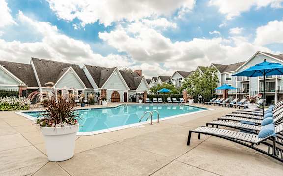 A pool surrounded by lounge chairs and umbrellas with a row of houses in the background at Huntington Cove Apartments, Merrillville, IN