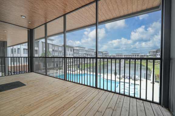 A wooden deck with a glass railing overlooking a pool and buildings.