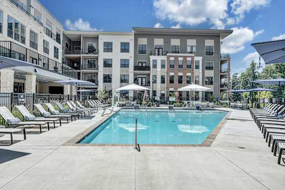 A large swimming pool surrounded by sun loungers and umbrellas in front of apartment buildings