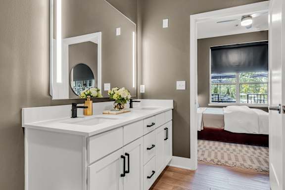 A bathroom with a vanity, mirror, and a bedroom in the background at Luxe 360 on Centerpointe Apartments, Virginia