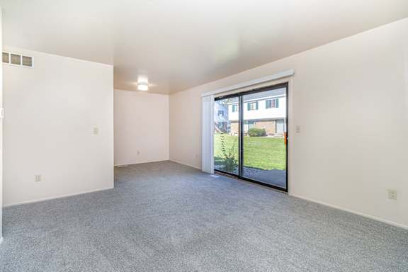 A large empty living room with a sliding glass door leading to a patio and green outdoor area at Mount Royal Townhomes in Kalamazoo, MI