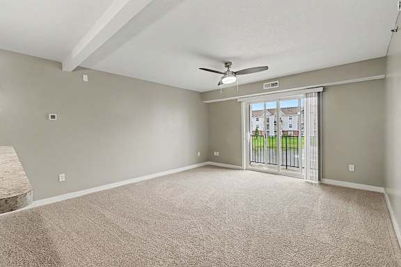 A living room with a carpeted floor and a ceiling fan leading to a balcony at The Reserve at The Reserve at Destination Pointe in Grimes, IA 50111