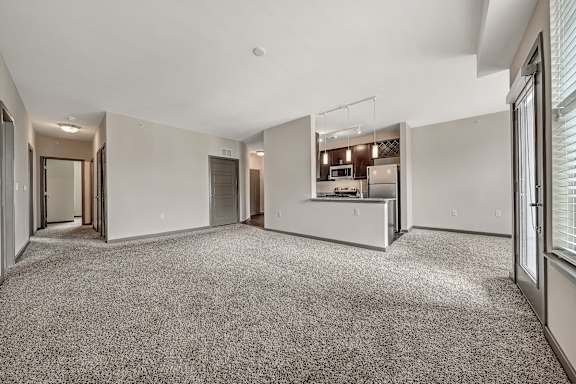 A spacious, empty room with a carpeted floor and a kitchen area in the background. at River Crossing Apartments, St. Charles, Missouri