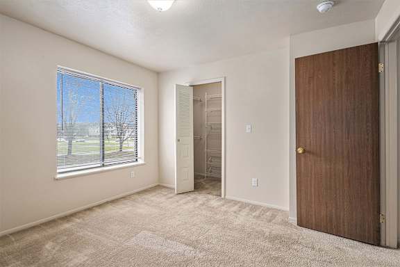 A room with a brown door, a window with a view of a tree-lined street, and a carpeted floor.