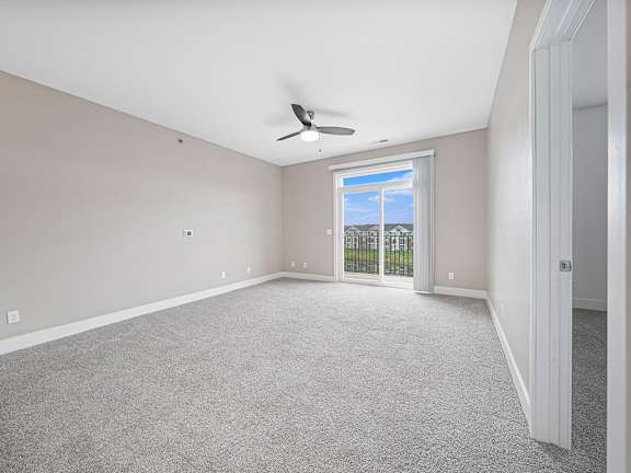 A living room with a ceiling fan and a window with a view of a fence and trees at Strathmore Apartment Homes in West Des Moines, IA