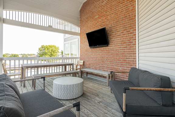 A balcony with a grey couch, a white ottoman and a flat screen TV mounted on a brick wall.