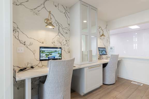 A white desk with a computer and chair in front of a cabinet with a wall hanging.
