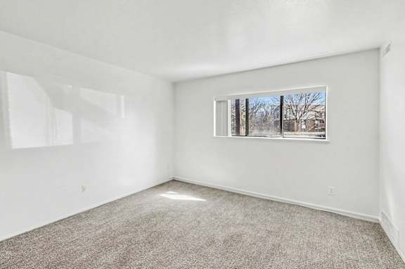 A living room with a carpeted floor and a window showing a view of trees outside at Trappers Cove Apartments, Lansing
