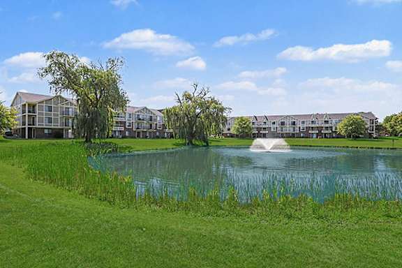 A serene landscape with a pond and a fountain in the foreground and apartment buildings in the background at Trappers Cove Apartments in Lansing, MI