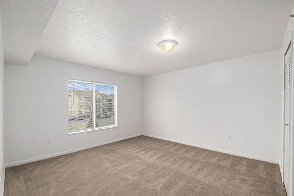 A large bedroom with a carpeted floor and a window showing a view of buildings outside at Trillium Pointe Apartment Homes, Michigan, 49201