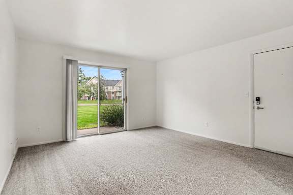 A living room with a carpeted floor and a sliding glass door leading to a patio and green lawn at Walnut Trail Apartments, Michigan