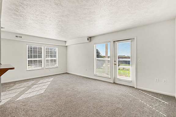 An enlarged living room with a carpeted floor, two windows with blinds, and a door leading to a patio at West Hampton Park Apartment Homes, Nebraska, 68022