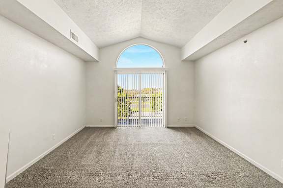 A living room with a cathedral ceiling, carpeted floor and a view of a balcony and trees through a window at West Hampton Park Apartment Homes, Nebraska, 68022