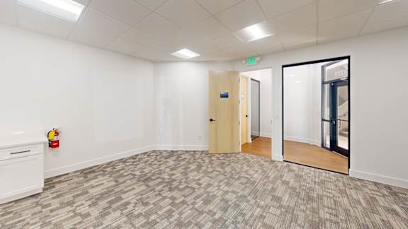 A room with a carpeted floor, a white wall, a wooden door, and a white cabinet with a red object on top.