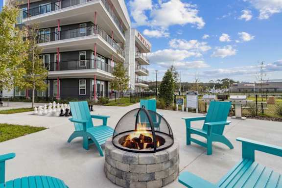 A fire pit sits in the middle of a patio with blue chairs and benches around it at Pinnacle Apartments, Jacksonville, 32256