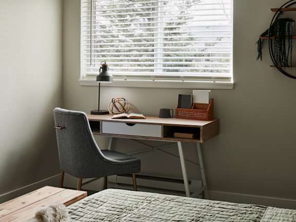 A grey chair sits in front of a desk with a lamp and some books on it.