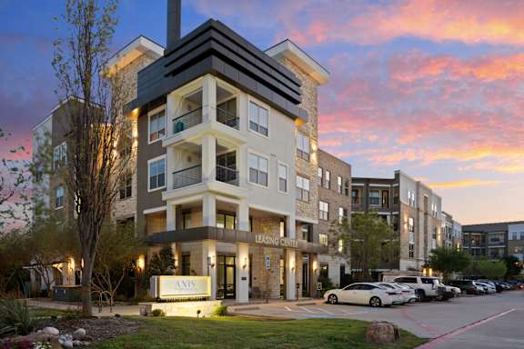 The image shows a modern apartment building with a parking lot in front of it.