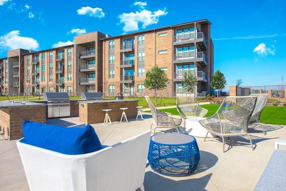A modern outdoor seating area with white chairs and blue cushions in front of apartment buildings.