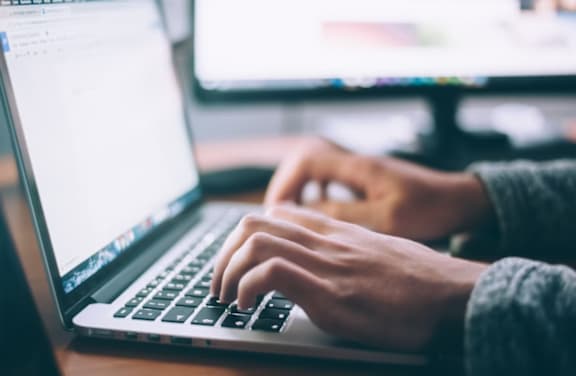 A person is typing on a laptop with a monitor in the background.