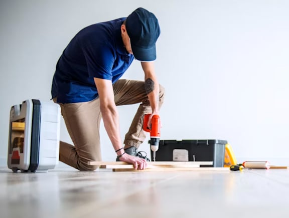 A man in a blue shirt and khaki pants is using a drill on a wooden plank.
