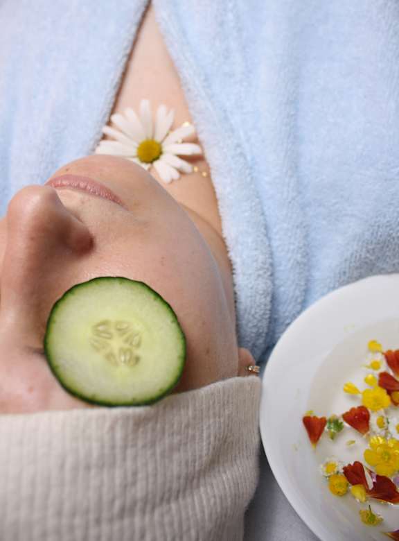 A person with a cucumber slice on their nose and a bowl of flowers nearby.